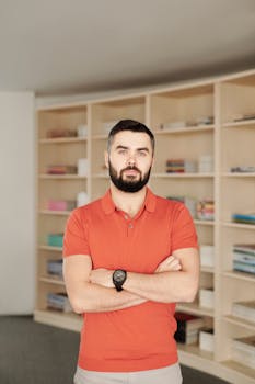 Portrait of a bearded man in a library wearing an orange polo, with books on wooden shelves in the background.